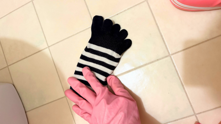 穴のあいたボーダー柄の五本指靴下を手袋越しに持ち、トイレ掃除に使おうとしている様子。床の上で撮影された写真。